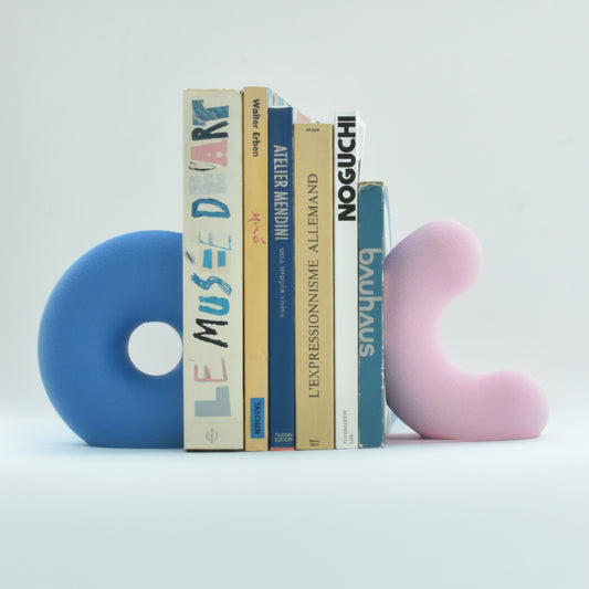 Une vue de présentation de deux serre-livres sculpturaux uniques de Benga Studio, tenant en place une collection de livres d'art et d'architecture. À gauche, un grand modèle bleu vif en forme de donut ouvert avec une texture mate. À droite, un modèle rose pastel en forme de vague sculpturale avec la même texture. Les deux sont maintiennent de gros et grand livres en place. Le bras de support discret sous chaque serre-livres permet de maintenir de gros livres. photo studio des objets sur un fond blanc.