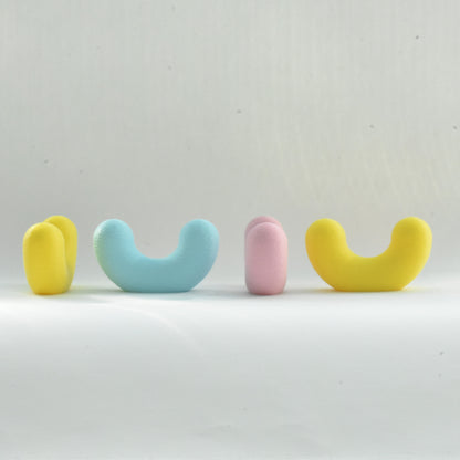 Quatre crochets muraux design en forme de U de la collection Benga Studio, alignés sur un fond blanc. De gauche à droite, la disposition permet de voir l'objet sous différents angles : un crochet jaune vu de profil, un bleu ciel vu de face, un rose pastel vu de profil, et un second crochet jaune vu de face. Ils présentent tous une finition mate et finement texturée, typique de l'impression 3D.