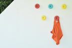 Mise en situation estivale de cinq crochets muraux design Benga Studio (modèle Donut) fixés sur un mur blanc en extérieur. Les patères rondes aux couleurs pop (rouge brique, bleu ciel, jaune vif) créent une composition murale ludique. Un maillot de bain une pièce orange est élégamment suspendu au crochet inférieur, illustrant parfaitement l'utilisation de ces patères résistantes comme accroche-serviettes ou porte-vêtements originaux aux abords d'une piscine.