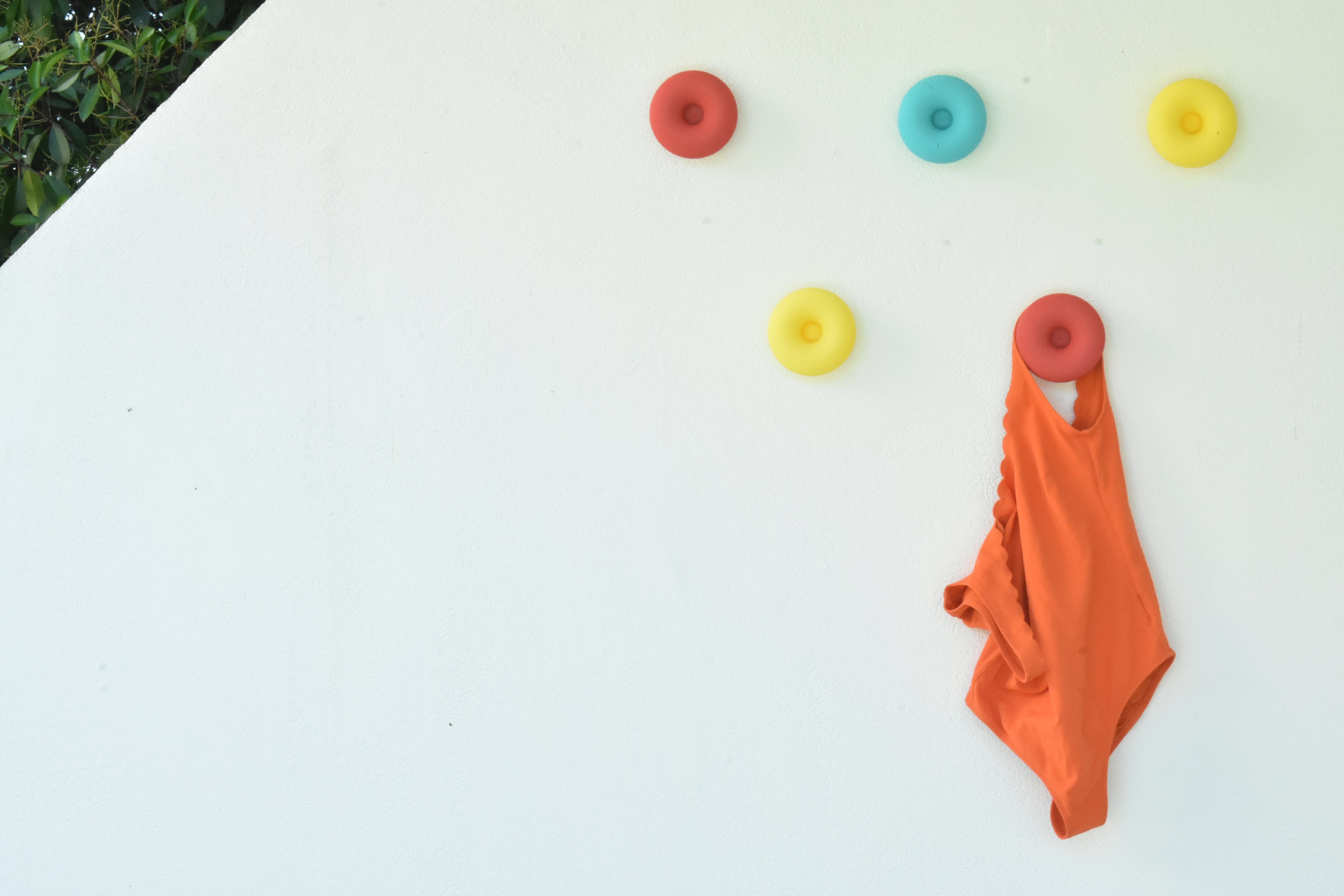 Mise en situation estivale de cinq crochets muraux design Benga Studio (modèle Donut) fixés sur un mur blanc en extérieur. Les patères rondes aux couleurs pop (rouge brique, bleu ciel, jaune vif) créent une composition murale ludique. Un maillot de bain une pièce orange est élégamment suspendu au crochet inférieur, illustrant parfaitement l'utilisation de ces patères résistantes comme accroche-serviettes ou porte-vêtements originaux aux abords d'une piscine.