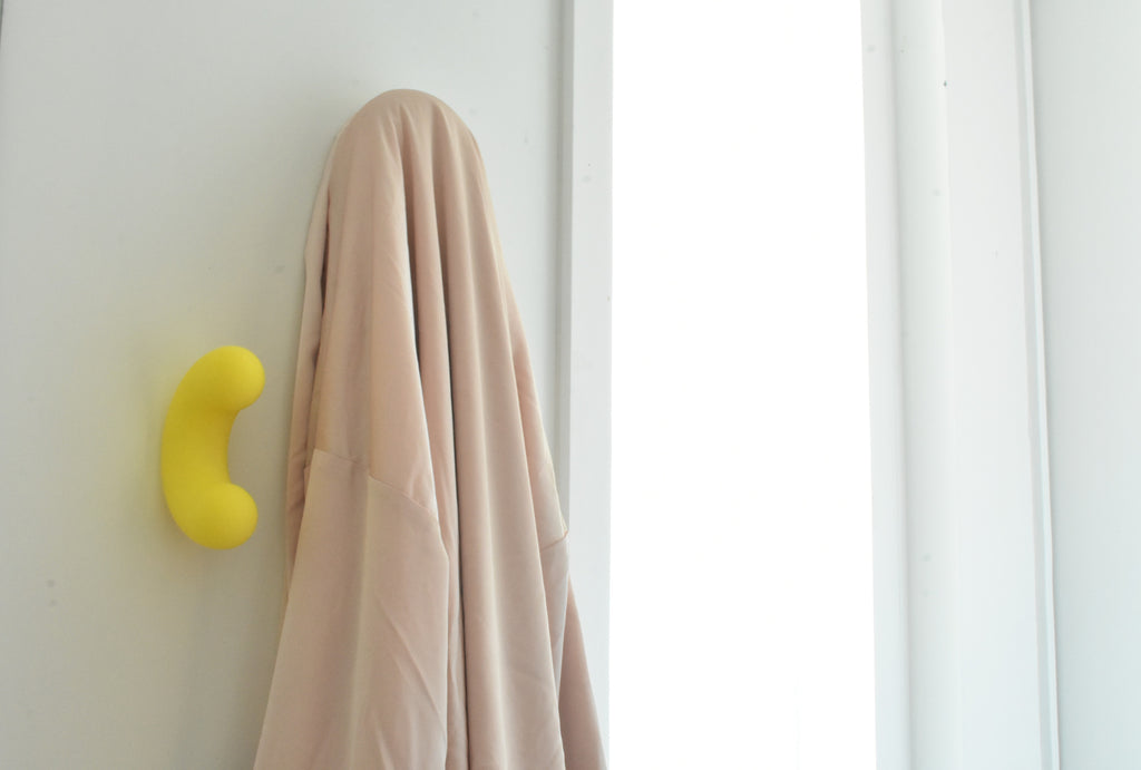 Mise en situation lumineuse d'un crochet mural design Benga Studio. Sur un mur blanc, près de l'encadrement d'une fenêtre, un crochet jaune vif en forme de U est solidement fixé. Juste à côté, un vêtement beige fluide est élégamment suspendu, illustrant parfaitement la fonction de porte-manteau de ces accroches décoratives et colorées au quotidien.
