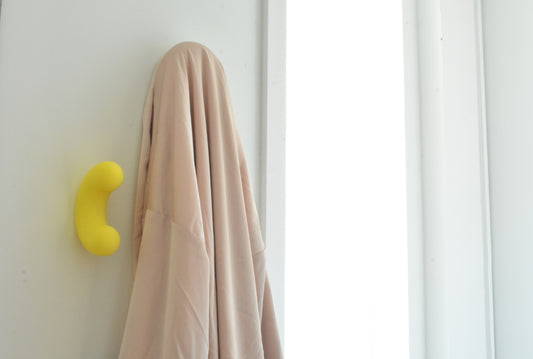 Mise en situation lumineuse d'un crochet mural design Benga Studio. Sur un mur blanc, près de l'encadrement d'une fenêtre, un crochet jaune vif en forme de U est solidement fixé. Juste à côté, un vêtement beige fluide est élégamment suspendu, illustrant parfaitement la fonction de porte-manteau de ces accroches décoratives et colorées au quotidien.