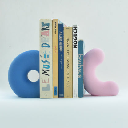 Une vue de présentation de deux serre-livres sculpturaux uniques de Benga Studio, tenant en place une collection de livres d'art et d'architecture. À gauche, un grand modèle bleu vif en forme de donut ouvert avec une texture mate. À droite, un modèle rose pastel en forme de vague sculpturale avec la même texture. Les deux sont maintiennent de gros et grand livres en place. Le bras de support discret sous chaque serre-livres permet de maintenir de gros livres. photo studio des objets sur un fond blanc.