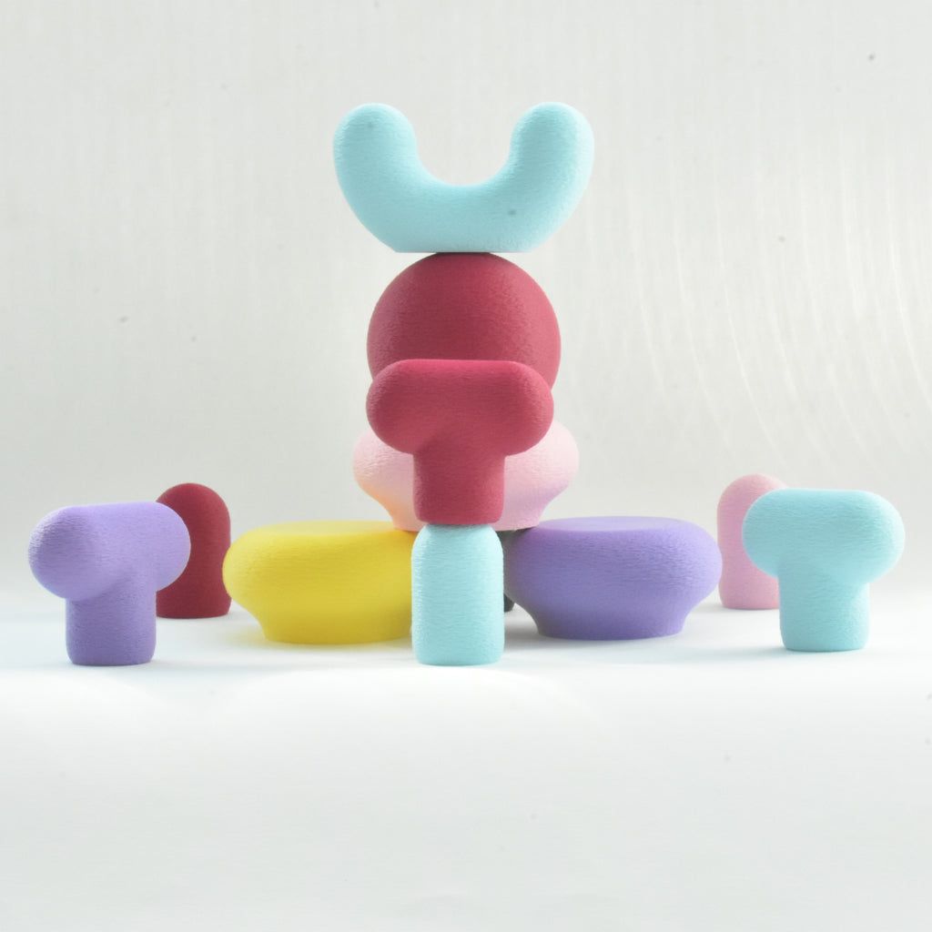 Une mise en scène studio sculpturale et ludique des crochets muraux colorés Benga Studio sur fond blanc. Au centre, plusieurs crochets mates sont empilés en équilibre à la manière d'un totem : un modèle bleu ciel en forme de I à la base, surmonté d'un crochet T bordeaux, d'un rond bordeaux, et couronné par un crochet en U bleu ciel. Autour de cette tour centrale sont parsemés d'autres crochets aux formes géométriques variées (T, galets, cylindres) dans des tons jaune vif, violet, rose pastel et bleu ciel. 