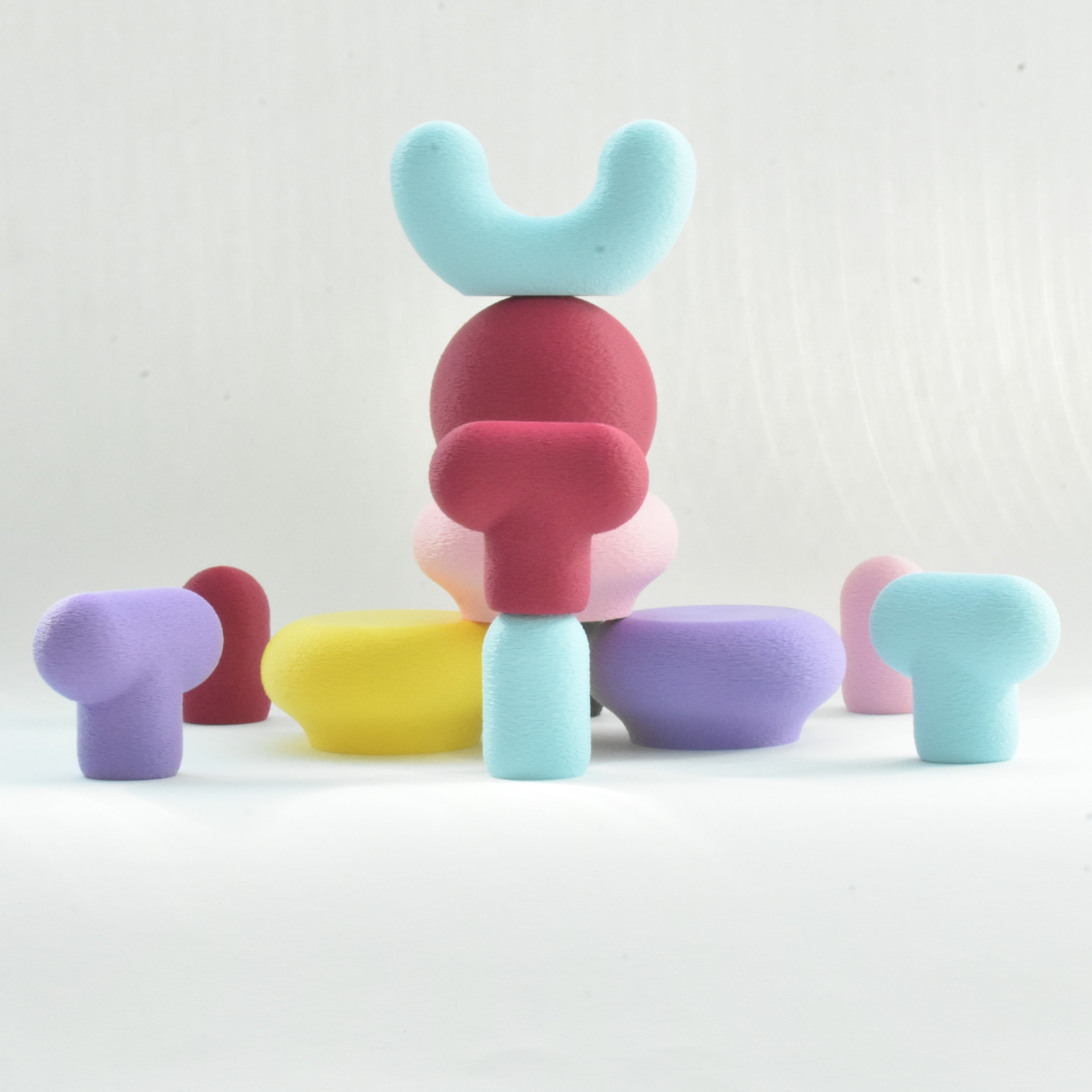 Une mise en scène studio sculpturale et ludique des crochets muraux colorés Benga Studio sur fond blanc. Au centre, plusieurs crochets mates sont empilés en équilibre à la manière d'un totem : un modèle bleu ciel en forme de I à la base, surmonté d'un crochet T bordeaux, d'un rond bordeaux, et couronné par un crochet en U bleu ciel. Autour de cette tour centrale sont parsemés d'autres crochets aux formes géométriques variées (T, galets, cylindres) dans des tons jaune vif, violet, rose pastel et bleu ciel. 